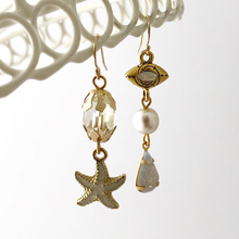 Load image into Gallery viewer, asymmetric earrings with starfish charm, vintage beads and evil eye connector hanging from a white frame