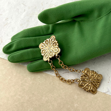 Load image into Gallery viewer, Gold floral cloak clasp or sweater clip with chain displayed on a green gloved hand on a mixed paper surface