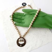 Load image into Gallery viewer, necklace with an an antique belt buckle and vintage champagne coloured pearl beads displayed on a gloved hand