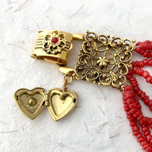 Load image into Gallery viewer, Gold heart-shaped locket and decorative clasp on red beaded bracelet against a white textured background