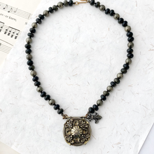 Load image into Gallery viewer, Necklace with black and gold beads and an ornate shield-like pendant on a textured white surface.