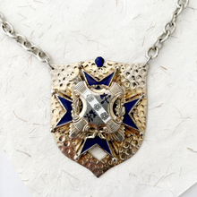 Load image into Gallery viewer, Close-up of necklace with a shield-shaped pendant featuring blue and gold design on a textured white background