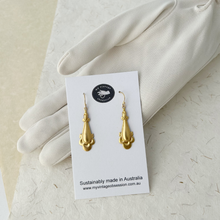 Load image into Gallery viewer, gold drop earrings with scrolled edges displayed on a white card resting against a gloved hand
