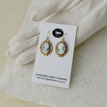 Load image into Gallery viewer, vintage blue resin cameo earrings in gold toned frames displayed on a white card resting against a gloved hand