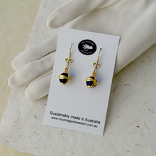 Load image into Gallery viewer, blue and gold orbs and gold plated fleur-de-lis connectors displayed on a white card leaning against a gloved hand