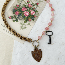 Load image into Gallery viewer, necklace with old medallion and key, cherry quartz beads and vintage chain displayed on a vintage postcard and paper
