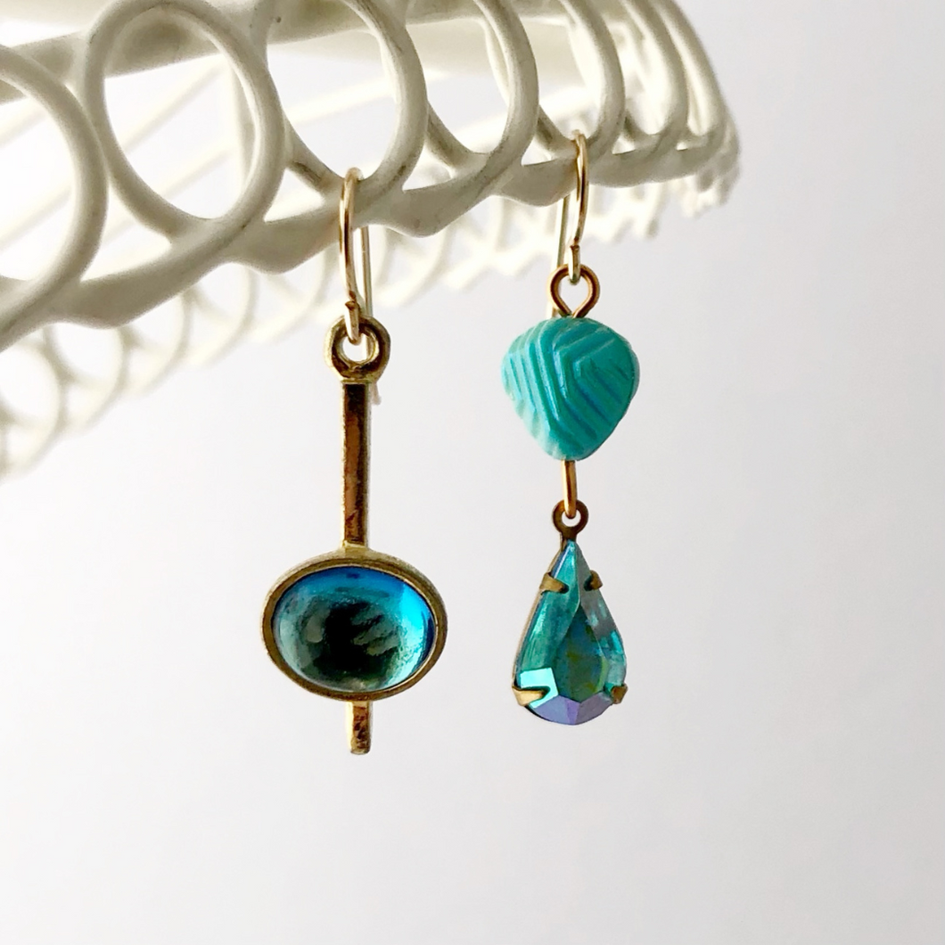 asymmetric earrings in iridescent aqua and gold hanging from a white frame