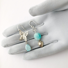 Load image into Gallery viewer, asymmetric earrings in aqua and silver tone with butterfly and heart charm displayed on a gloved hand
