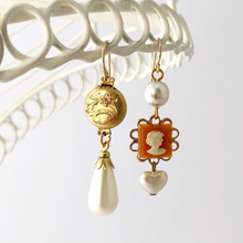 Load image into Gallery viewer, asymmetric earrings with gold filled locket, cameo, vintage pearl beads hanging from a white wire frame
