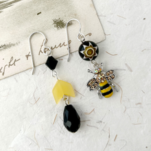 Load image into Gallery viewer, A pair of handmade earrings displayed on a textured white surface with a decorative card. They feature silver ear-wires, beads in yellow and black with a bee charm with rhinestone wings.
