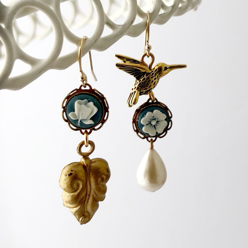 Pair of earrings with a gold hummingbird design, blue floral cameos, and a pearl drop on a white background.