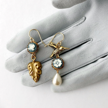 Load image into Gallery viewer, Pair of earrings with a gold hummingbird design, blue floral cameos, and a pearl drop on a grey gloved hand
