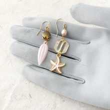 Load image into Gallery viewer, asymmetric earrings with shell, starfish, Mother of pearl buckle, pearl and pink leaf drop displayed on a grey gloved hand
