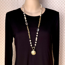 Load image into Gallery viewer, A long gold-tone and white pearl necklace with clear rhinestone accents and a round gold locket, displayed on a mannequin wearing a black long-sleeve top against a patterned beige wall.
