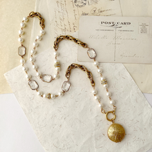 Load image into Gallery viewer, A sautoir-style pearl and gold necklace with rectangular clear beads and a gold locket displayed on textured paper next to vintage postcards.
