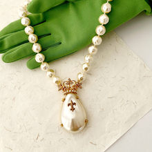 Load image into Gallery viewer, Faux pearl necklace with a decorative crowned pendant on a textured surface on a green gloved hand.
