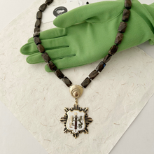 Load image into Gallery viewer, Necklace with an enamelled heraldic pendant displayed on a green-gloved hand on a textured white surface.
