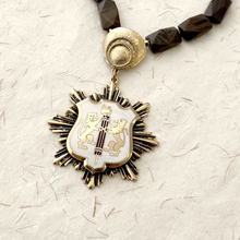 Load image into Gallery viewer, Close-up of vintage enamelled heraldic pendant in necklace against a textured paper background
