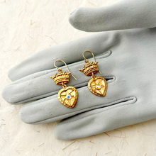 Load image into Gallery viewer, Gold heart-shaped earrings with crowns on a gray glove.
