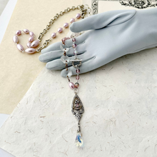 Load image into Gallery viewer, A reimagined vintage necklace with pink pearls, chandelier crystals,rhinestone filigree pendant and clear crystal drop, displayed on a grey gloved hand over textured papers.
