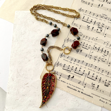 Load image into Gallery viewer, A vintage-style gold chain necklace with faceted dark red beads and a large leaf shaped pendant featuring red and green floral inlay, styled on antique sheet music.

