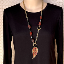 Load image into Gallery viewer, A long gold-tone chain necklace with red and brown stone beads and a large leaf-shaped pendant, displayed on a mannequin wearing a black long-sleeve top.
