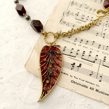 Load image into Gallery viewer, A close-up of a gold-tone pendant necklace with a red enamel leaf-shaped charm decorated with small multicolored floral details, shown resting on vintage sheet music with dark red beads on a white textured surface.
