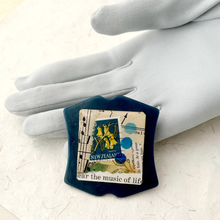 Load image into Gallery viewer, A blue handmade brooch with a collage featuring vintage paper and a New Zealand stamp, displayed against a grey gloved hand.

