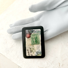 Load image into Gallery viewer, A rectangular black-framed collage of vintage illustrations, including a green Australia stamp, botanical drawings, and musical notes, displayed on a white textured surface with a grey gloved hand behind it.
