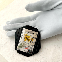 Load image into Gallery viewer, A black octagonal ring featuring a vintage botanical and musical collage design, displayed on a white textured surface next to a grey glove.

