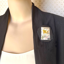 Load image into Gallery viewer, A close-up of a mannequin torso wearing a black blazer over a white top, with a decorative rectangular pin or brooch pinned to the left lapel. The pin has a collage design with a yellow stamp, green leaves, and text elements.
