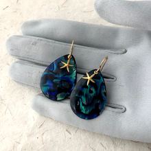 Load image into Gallery viewer, Teardrop-shaped paua shell earrings with starfish designs on a grey gloved hand.
