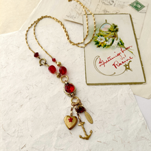 Load image into Gallery viewer, A gold-toned long necklace with red beads, a large red gem, a small heart charm, a peace charm and an anchor charm, displayed on textured white paper next to a vintage post card.
