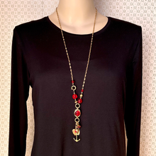 Load image into Gallery viewer, A long gold-tone necklace with red beads and a dangling anchor charm with a small heart detail, displayed on a mannequin wearing a black long-sleeve top against a patterned beige background.
