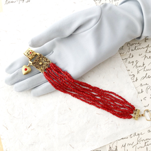 Load image into Gallery viewer, Red beaded bracelet with gold clasp on a gray gloved hand against a textured surface.
