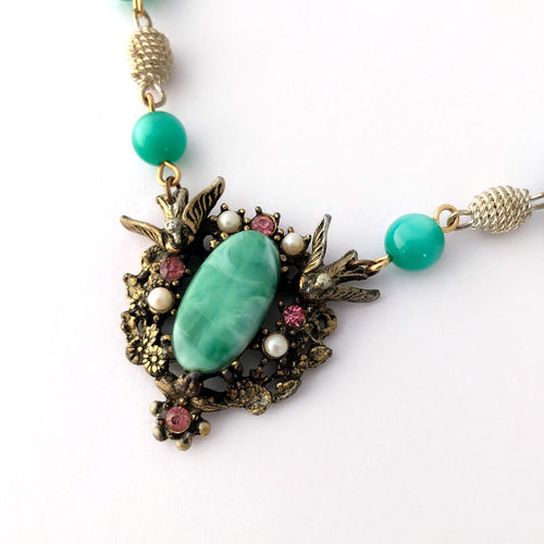 close up of necklace focal with two birds, pink rhinestones and pearls surrounding a large green oval