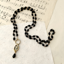 Load image into Gallery viewer, A beaded necklace with faceted black beads and gold-toned metal accents, featuring a vintage oval pendant with a portrait and a teardrop drop. The necklace is arranged on a textured white sheet of paper over a sheet of printed music.

