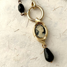 Load image into Gallery viewer, A close-up of a gold-toned necklace featuring a classic oval cameo pendant with a black and white profile design, accented by a dangling black teardrop bead and a small black faceted bead on the chain, set against a textured off-white background.
