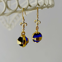 Load image into Gallery viewer, earrings made with blue and gold orbs and gold plated fleur-de-lis connectors hanging from a white wire frame
