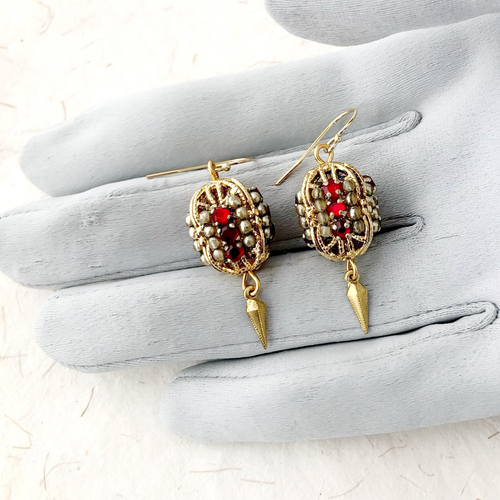 Gold bead earrings with red stones and glass pearls displayed on a grey gloved hand.